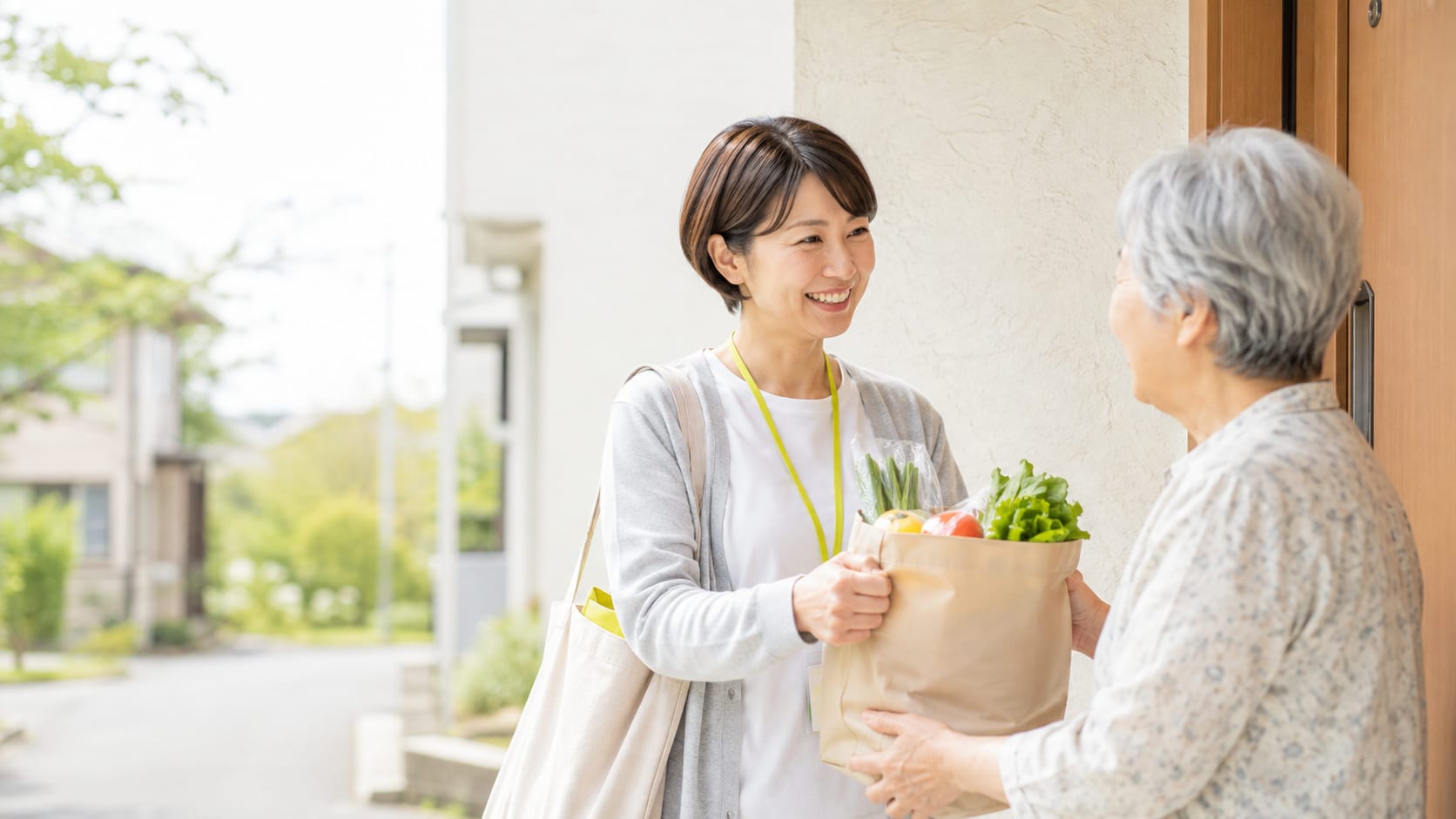 住宅街の玄関先で、買い物代行のスタッフが野菜の入った紙袋を高齢の女性に手渡し、笑顔で言葉を交わしている様子